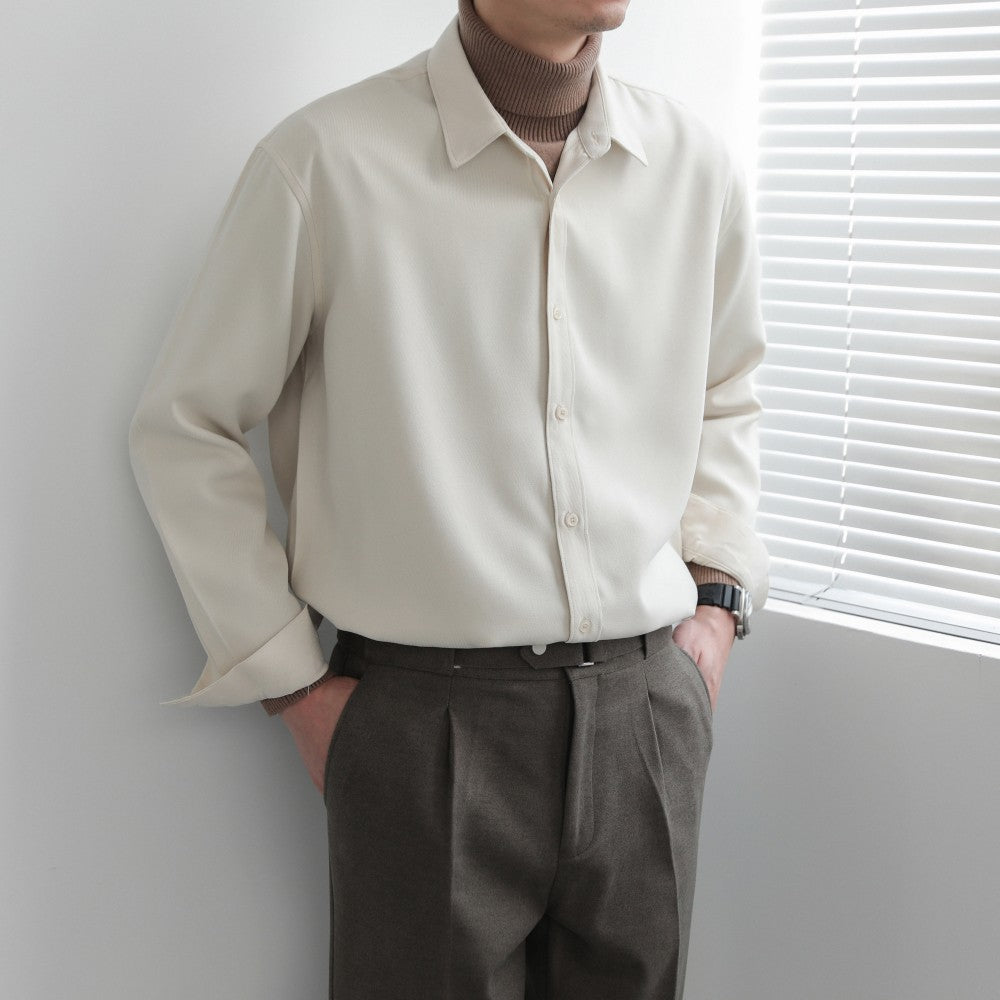 Person wearing a beige shirt and brown pants standing against a light gray wall with a window with blinds.